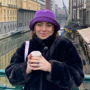 Portretfoto van Aisling, oud-deelnemer Futureproof. Ze staat op een brug met gebouwen op de achtergrond. Ze houdt een koffiebeker vast en draagt een paarse bucket hat.